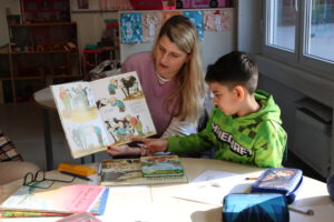 Eine Frau in einem lila Pullover hält ein geöffnetes Bilderbuch mit Kuh-Illustrationen hoch und liest zwei Kindern an einem Tisch vor.