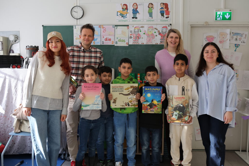 Gruppenbild vor einer Schultafel: Nicolas Berthold und Ann-Katrin Herke posieren gemeinsam mit den Übungsleitungen von Chancenwerk und einer Gruppe von Kindern, die stolz ihre Bilderbücher zeigen.