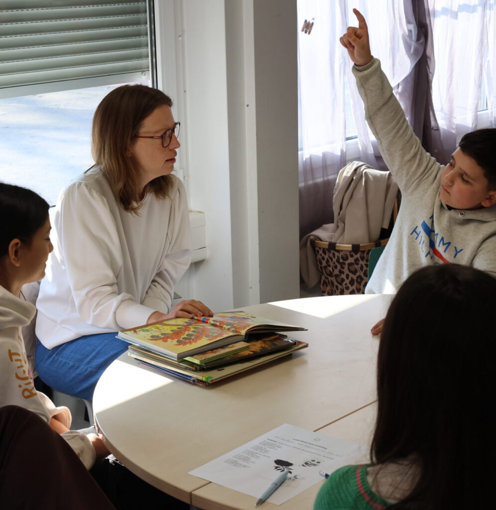 In einer kleinen Gruppe am Tisch meldet sich ein Junge mit erhobenem Zeigefinger, während Meira Schmidt von der Postcode Lotterie mit Brille vor einem Stapel Bilderbücher sitzt und ihm zuhört.