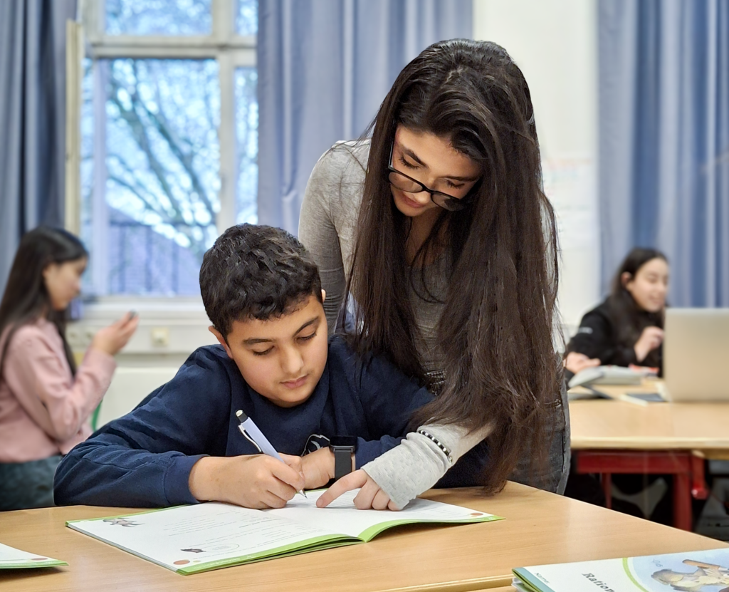 Eine Betreuerin mit Brille beugt sich über einen Schüler und zeigt auf eine Aufgabe in seinem Heft, während er schreibt. Im Hintergrund sind weitere Jugendliche im Klassenzimmer zu sehen.