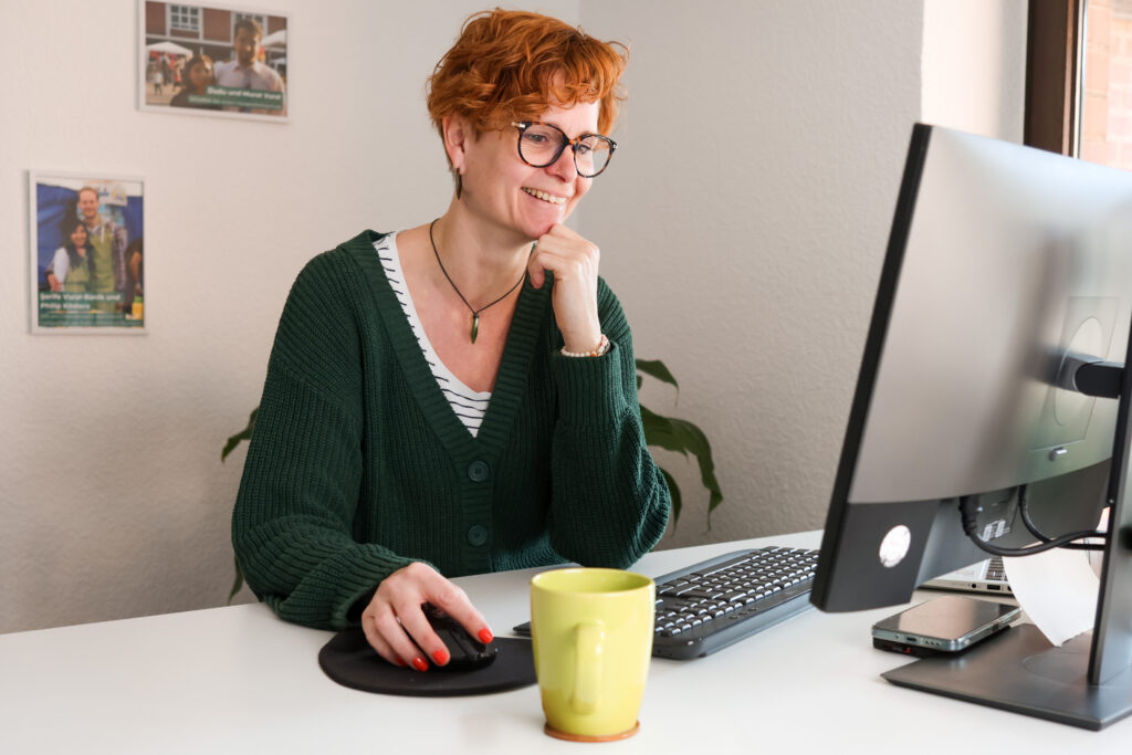 Jasmin Brozulat mit kurzen roten Haaren und Brille sitzt lächelnd an einem Schreibtisch und arbeitet an einem PC-Monitor.